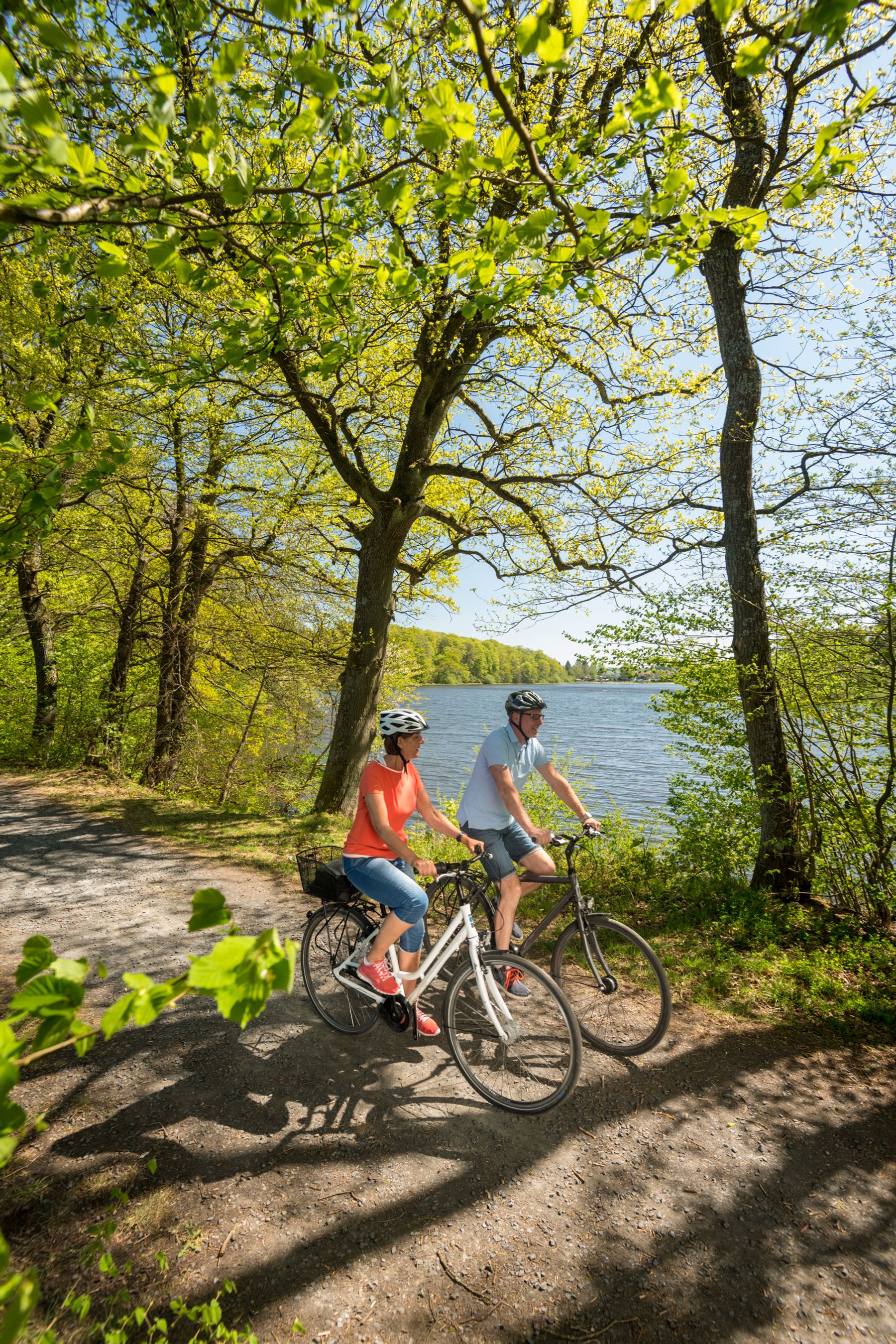 Zwei Personen mit Helmen fahren mit dem Fahrrad auf einem sonnenbeschienenen, von Bäumen gesäumten Weg neben einem See. Die Szene ist hell und grün, mit Frühlingsblättern und klarem, blauem Wasser im Hintergrund.
